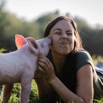 Schwein Anni (als junges Ferkel) berührt mit ihrem Rüssel eine weiblich gelesene Person an der Wange, die auf dem Boden im grünen Gras liegt und Anni mit den Händen am Hals berührt und streichelt. Die Person hat geschlossene Augen und verzieht verschmitzt das Gesicht. Im Hintergrund sind verschwommen Bäume zu erkennen.