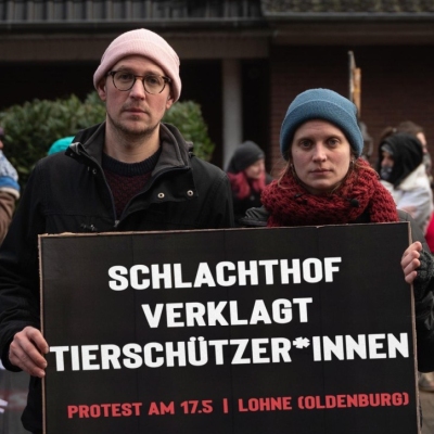 Zwei Aktivist*innen halten ein Schild mit der Aufschrift "Schlachthof verklagt Tierschützer*innen. Protest am 17.5. | Lohne (Oldenburg)"