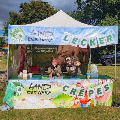 Zwei Menschen an einem bunten Infostand vom Land der Tiere mit der Aufschrift "Lecker Crepes".
