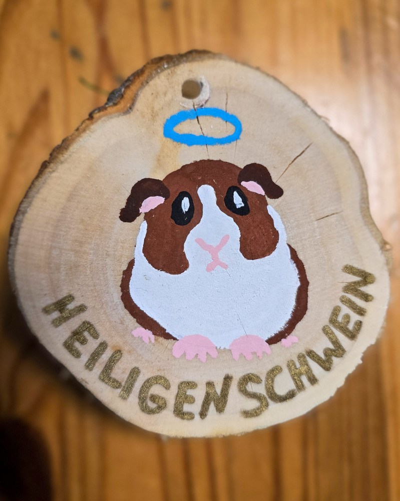 Eine bemalte Deko.Holzscheibe mit einem Meerschweinchen mit Heiligenschein. Dazu der Text "Heiligenschwein"