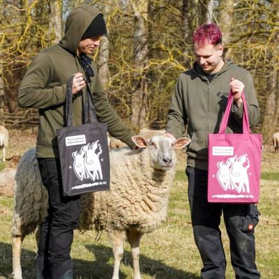Zwei Menschen mit dunkelgrauer bzw. himbeer-farbenen Tragetaschen und dazwischen Schaf Biene